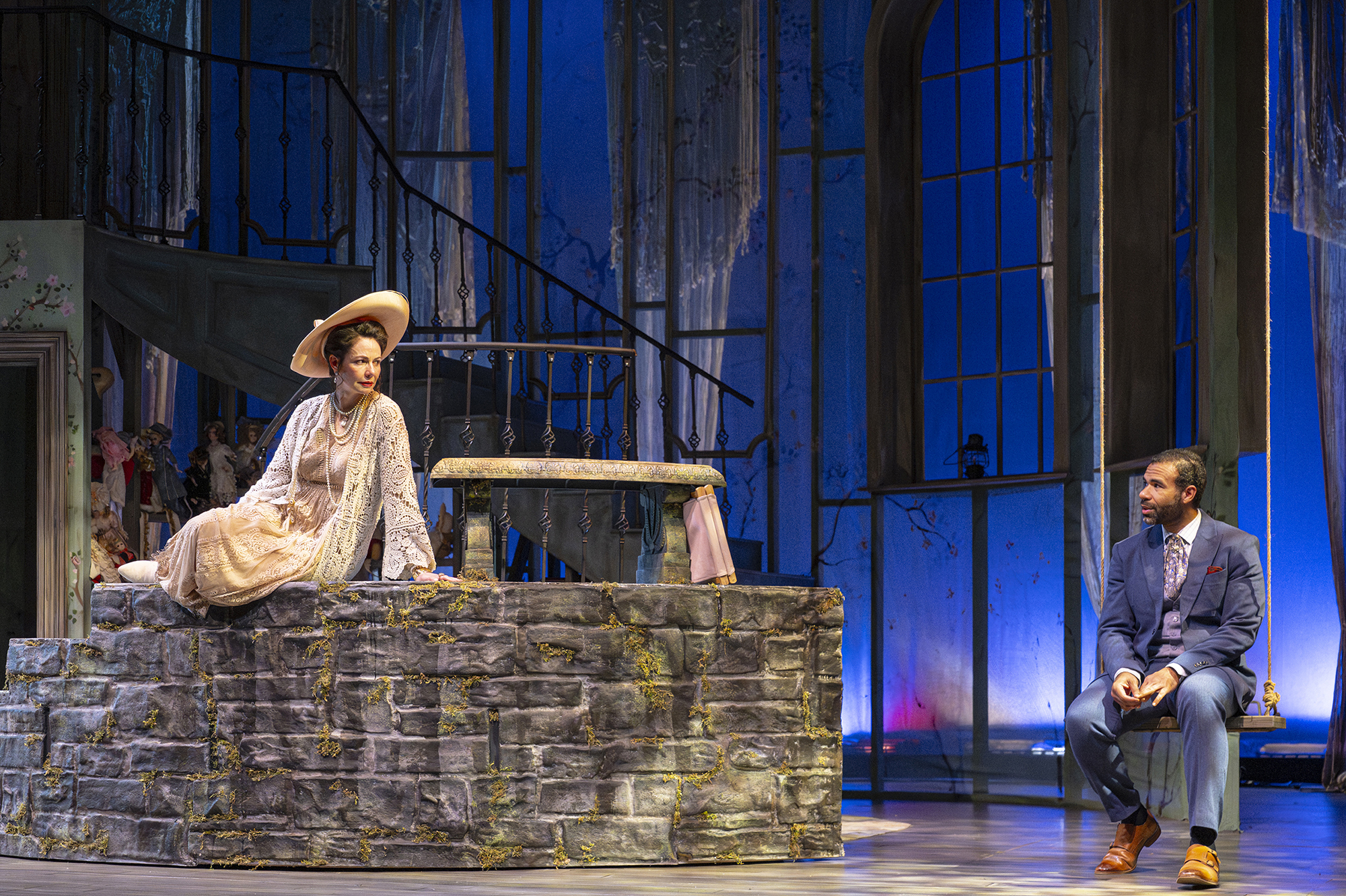 A white woman in a dress and large hat sits on a stone wall, looking to the right at a Black man in a blue suit, sitting on a swing
