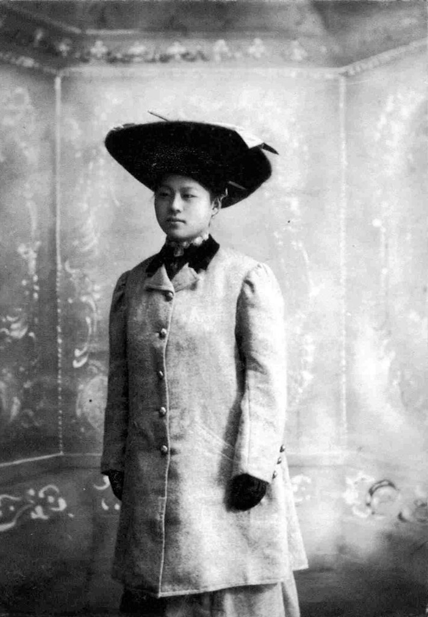 An early 20th century portrait of a Chinese woman wearing a long formal coat and a black hat.