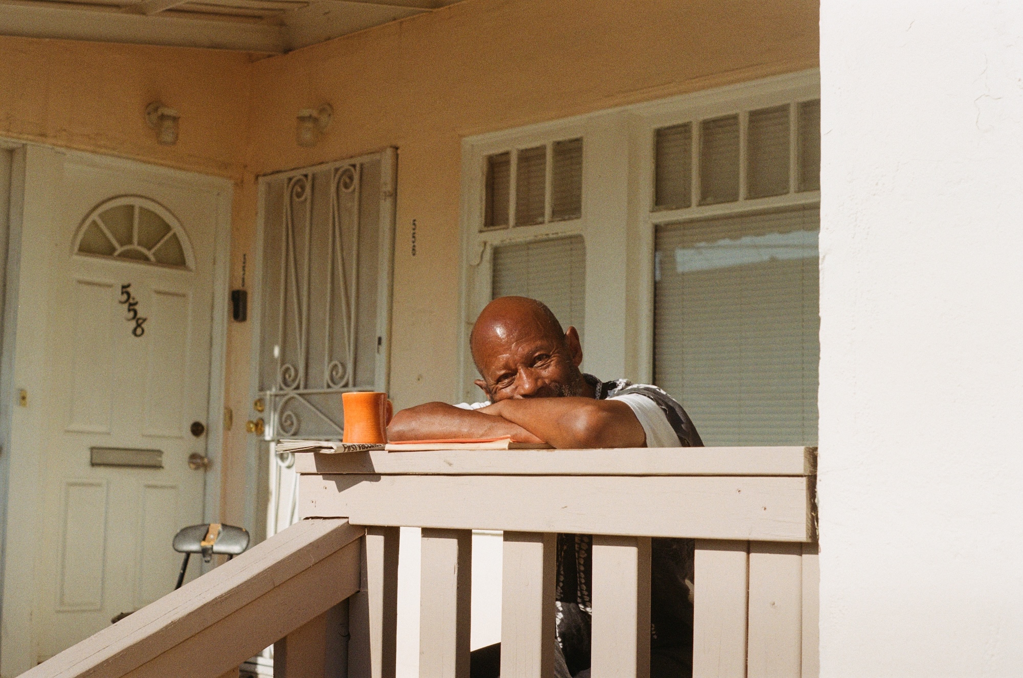 An elder Black man rests his arms on a handrail as he sits on the front porch of a house. 