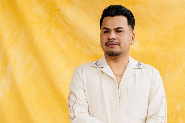 A man in a white suit with patterned decor stands against a yellow backdrop.