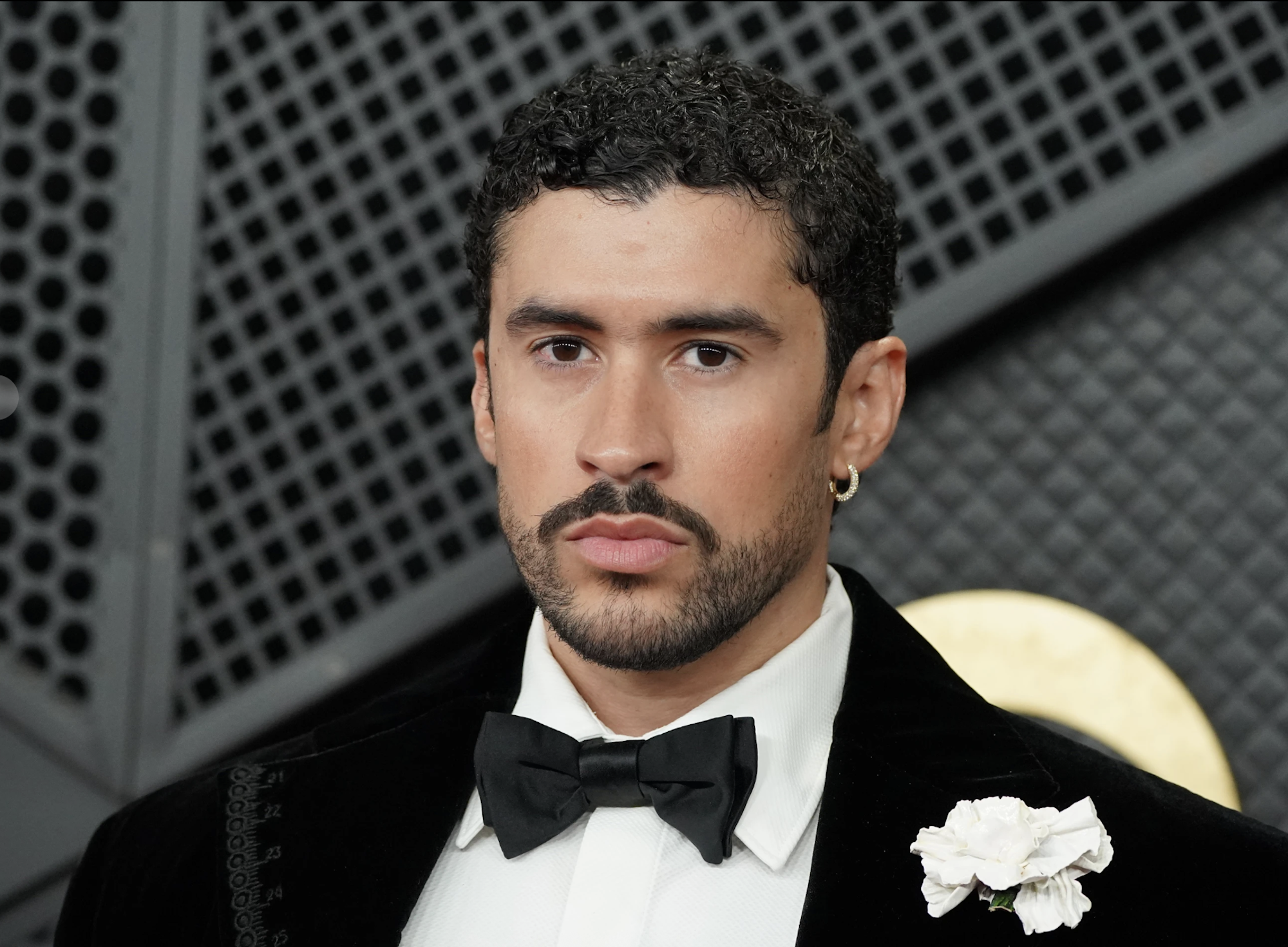 A Latino man with groomed beard and neatly curled hair wears a tuxedo on a red carpet.