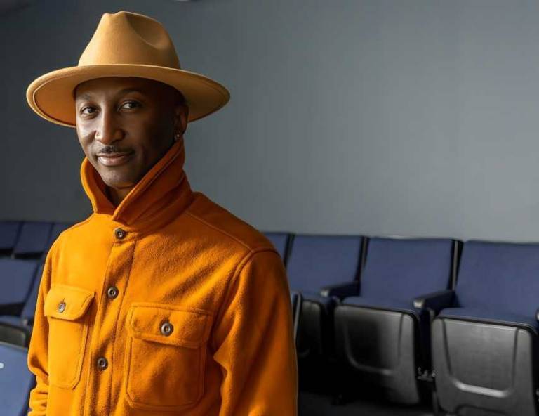 An African American man in a stylish orange hat and coat poses for a photo while standing in front of a row of chairs inside of an auditorium.