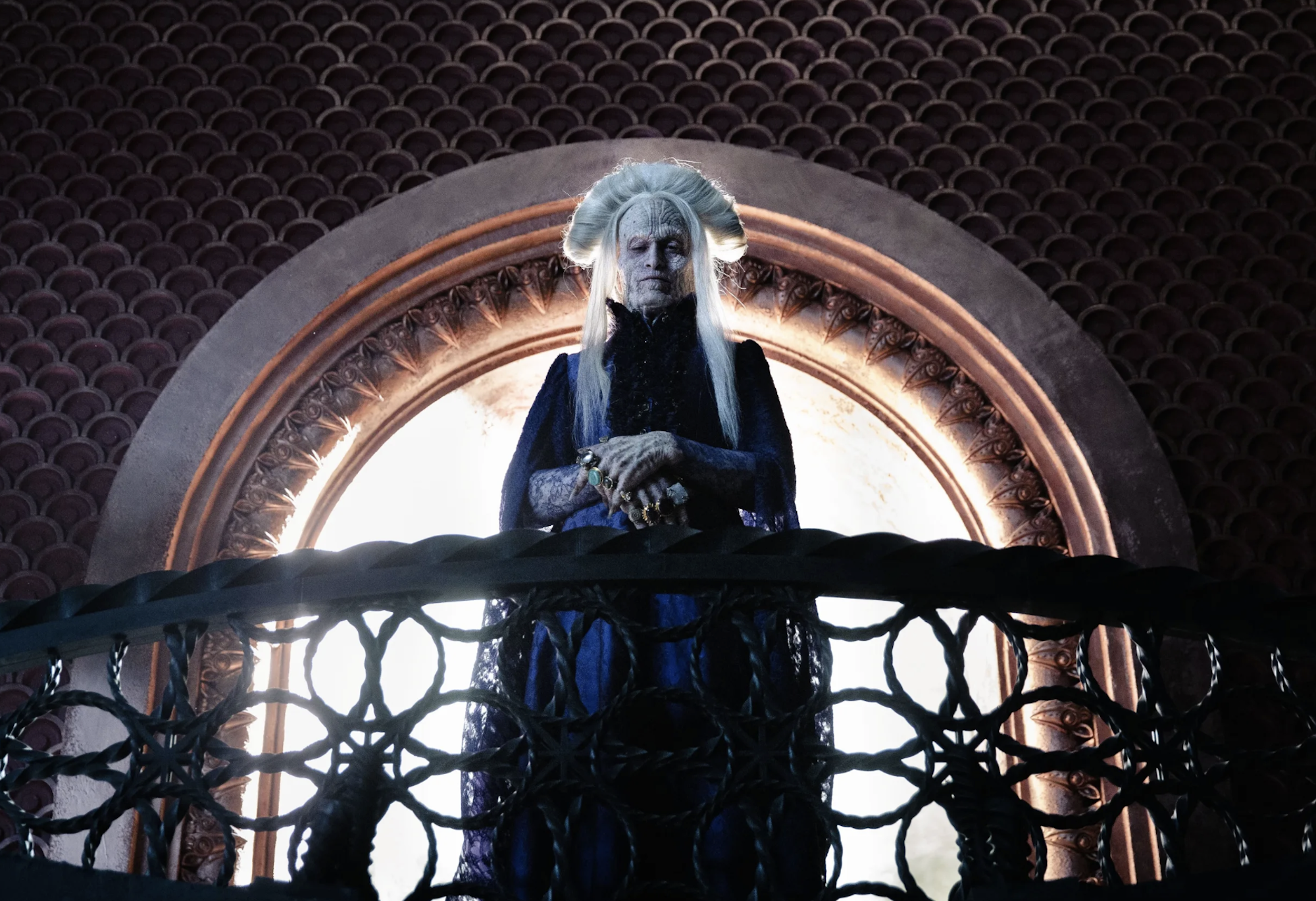A gothic figure wearing a long dark cape stands on a balcony, backlit from the room behind him. His face is deathly white and aged, and his hair is long and white.