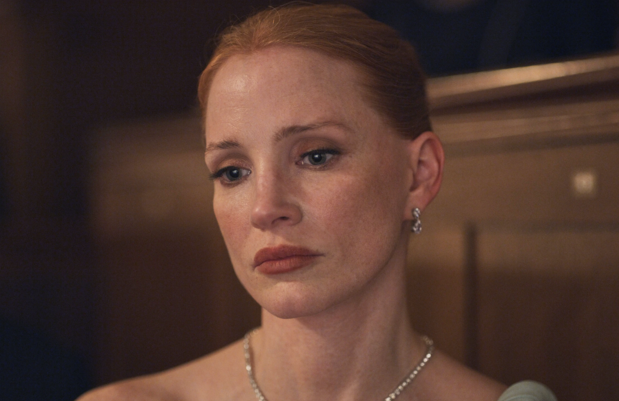 A well groomed white woman wearing diamond jewelry sits looking glum in a theater box.