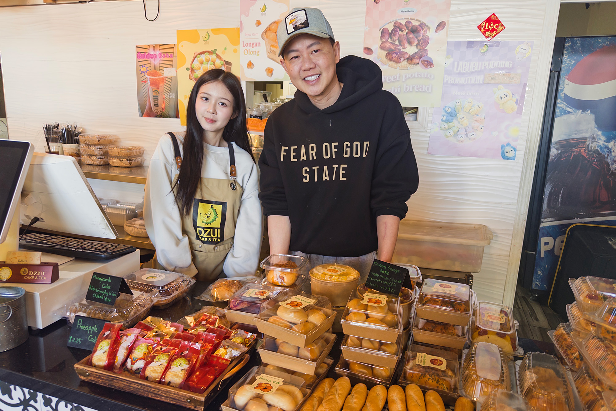 A man and a young woman of Asian descent pose in front of a display of cakes and pastries.