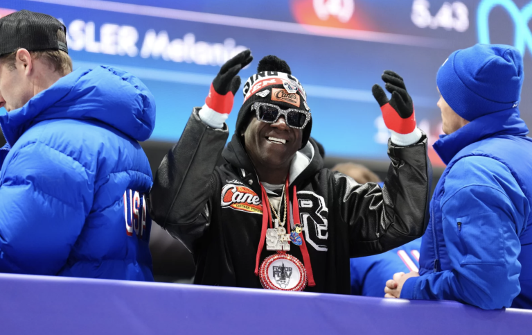 A Black man wearing winter clothes and sunglasses smiles and gestures from a balcony inside a sports arena.