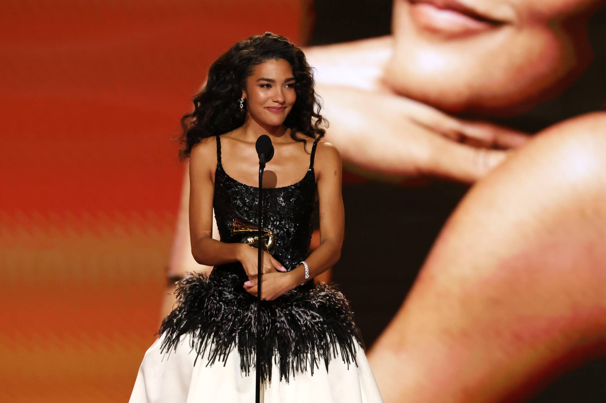 A slender attractive Black woman with long wavy hair, wearing a black and white gown, accepts a Grammy award on stage.