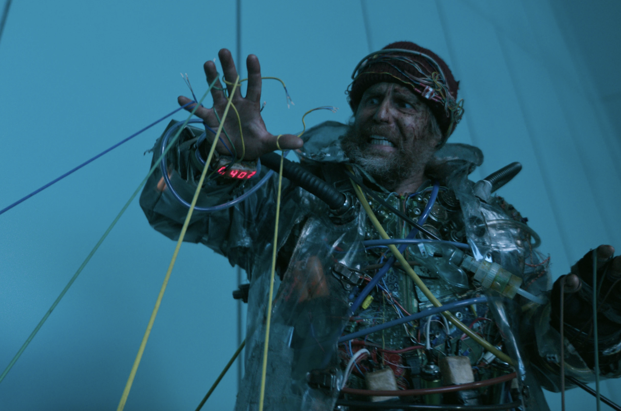 A bearded grimacing man examines one hand inside a stark blue room. He is wearing an outfit made of wires and garbage.