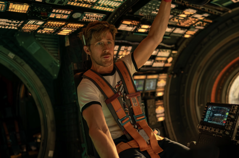 A middle aged man in a white t shirt sits strapped into a seat inside a space ship.