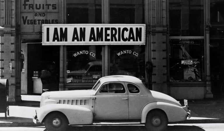 A black and white image of a 1940s-era store, with white car parked outside. A large sign reading 'I am an American' hangs on the store front.