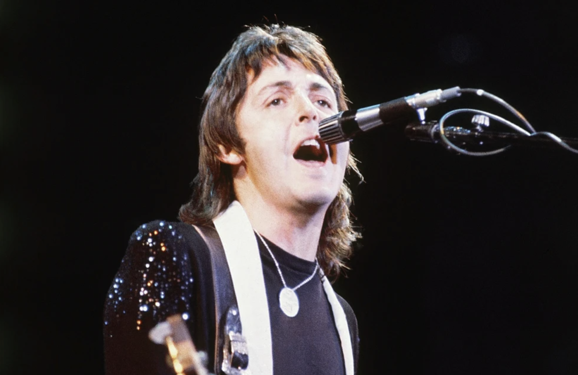1970s-era Paul McCartney performing on stage, singing into a microphone.