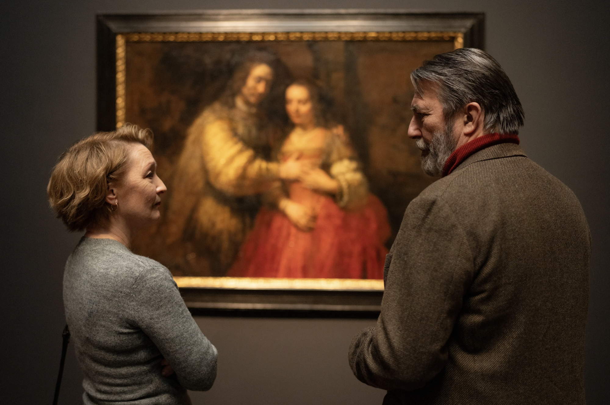 An older white couple stand in a gallery, looking at each other as they stand in front of a renaissance painting.