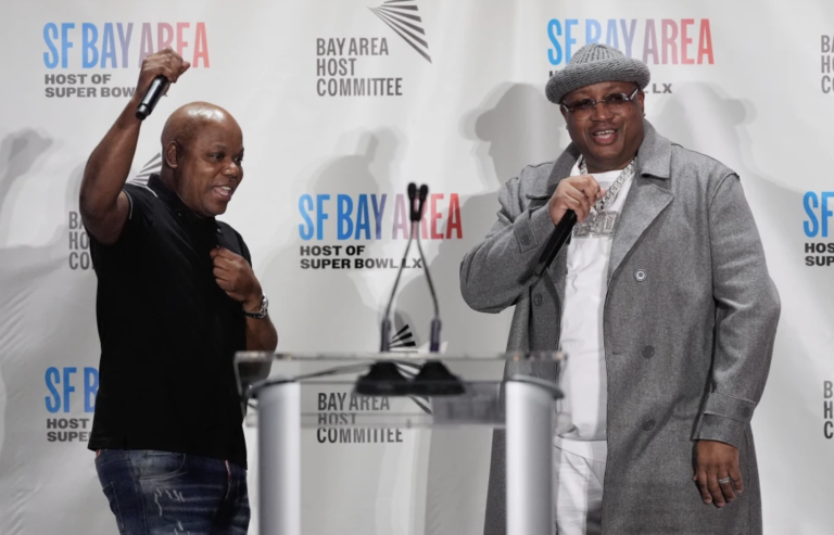 A bald Black man cheers with one fist in the air while at a podium on stage at a press conference. To his left is a Black man wearing a grey overcoat and matching hat. Both are smiling.