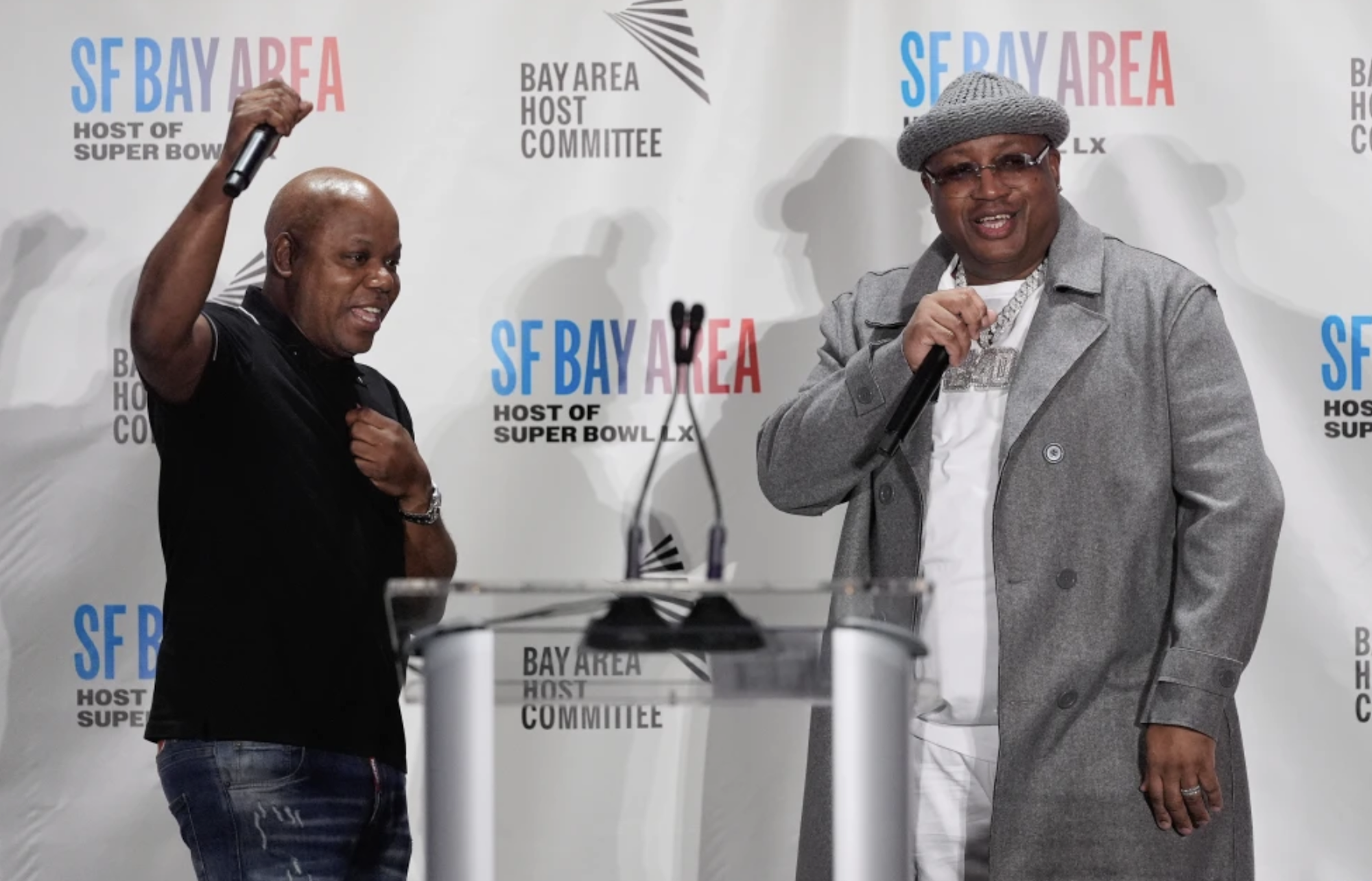 A bald Black man cheers with one fist in the air while at a podium on stage at a press conference. To his left is a Black man wearing a grey overcoat and matching hat. Both are smiling.