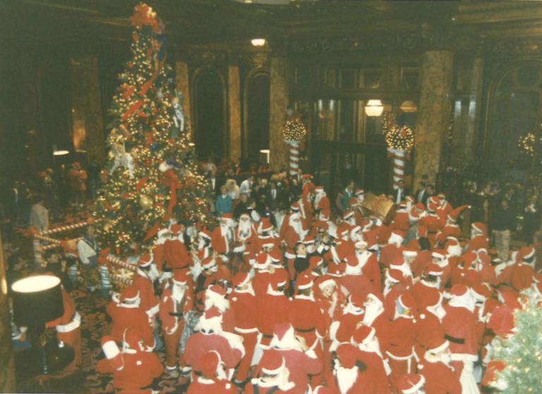 A luxury hotel lobby is seen from above, filled almost entirely with people in red and white Santa suits. A large Christmas tree is also inside.