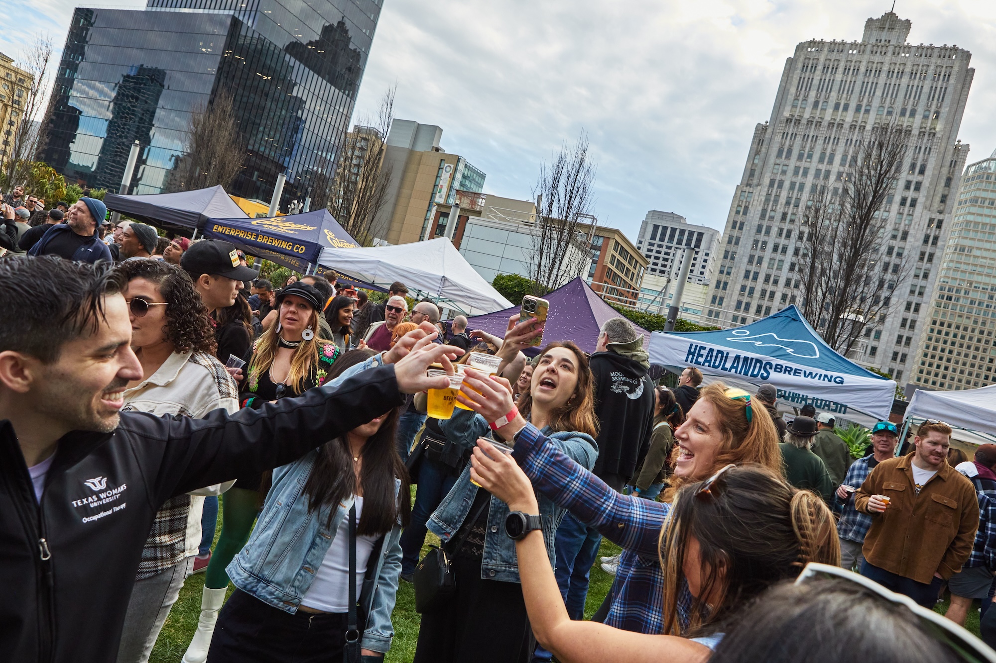 People gathered in a circle, standing outside and raising their glasses as they cheers with beer.