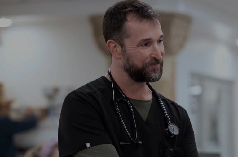 A white, male, middle-aged doctor wearing black scrubs with a stethoscope around his neck.