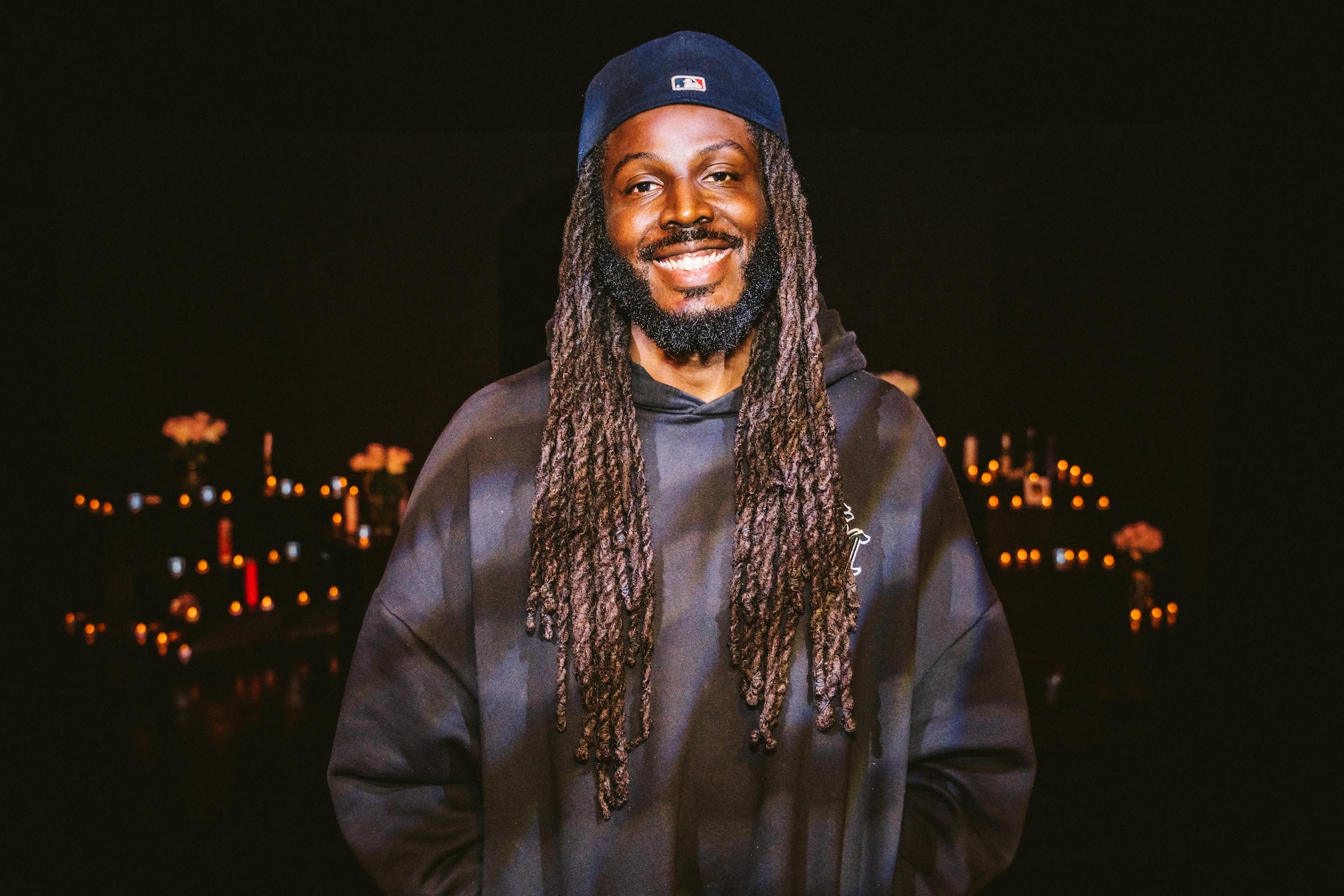 An African-American man with long locs and a backwards baseball hat wears a blue and black hoodie as he smiles for a photo. 