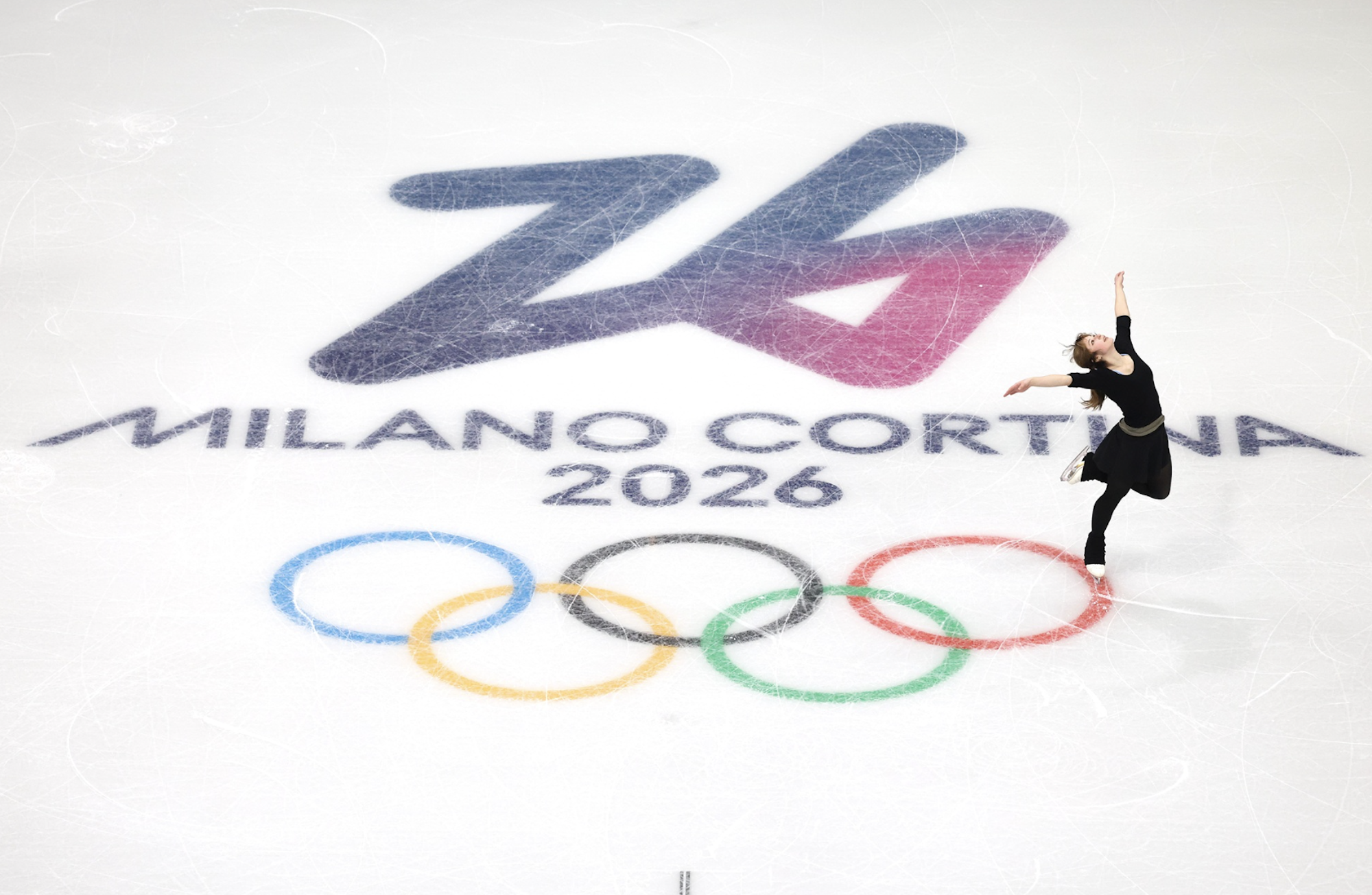 An iceskater in black strikes a pose at the edge of olympic rings depicted on the ice.