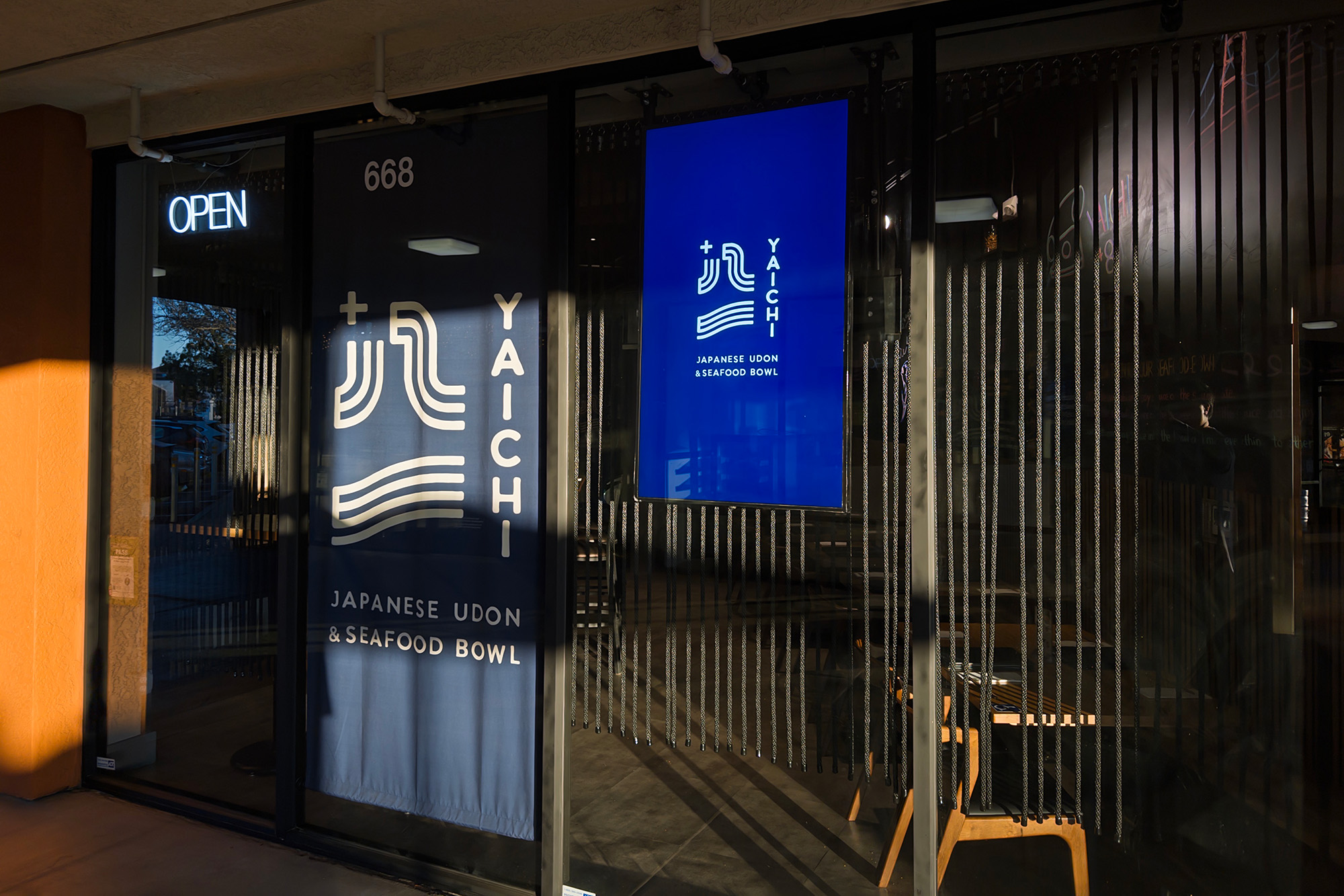 Exterior of a restaurant. The blue curtain hanging in the doorway reads, "Yaichi Japanese Udon & Seafood Bowl."
