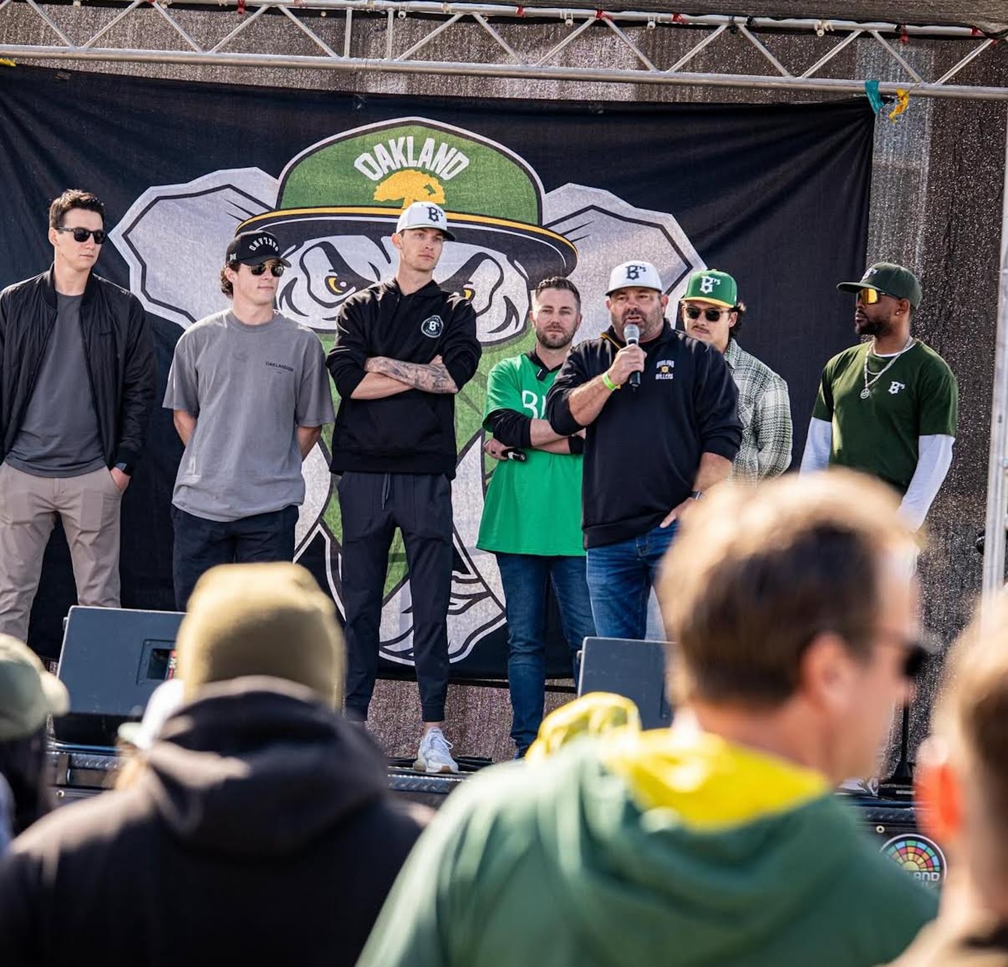 Seven men stand on stage, one holding a microphone, addressing a crowd at a festival.