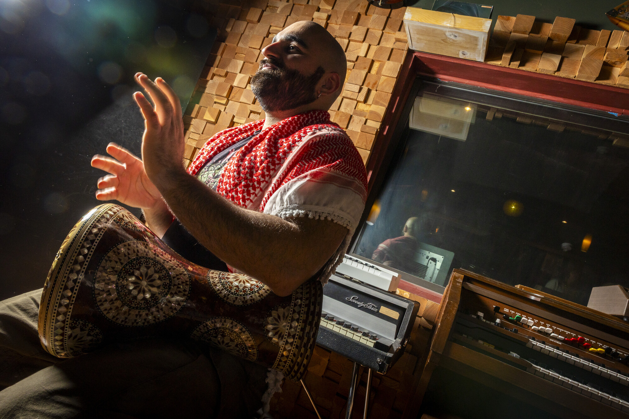 A man in a keffiyeh scarf plays a hand drum in a recording studio.