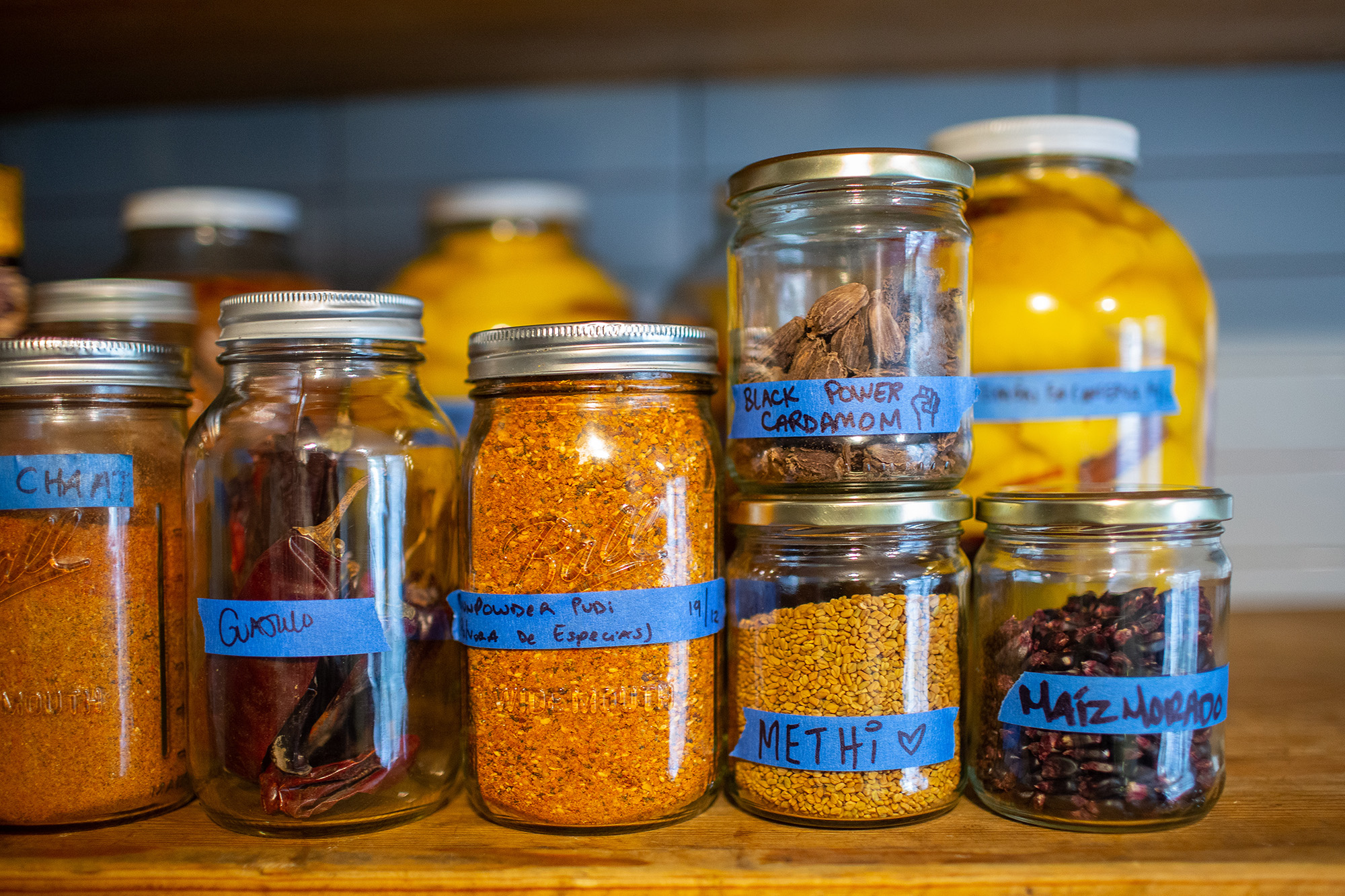 Jars of spices labeled with blue tape, including dried guajillo chiles, gunpowder pudi, "Black Power cardamom," methi, and maiz morado.