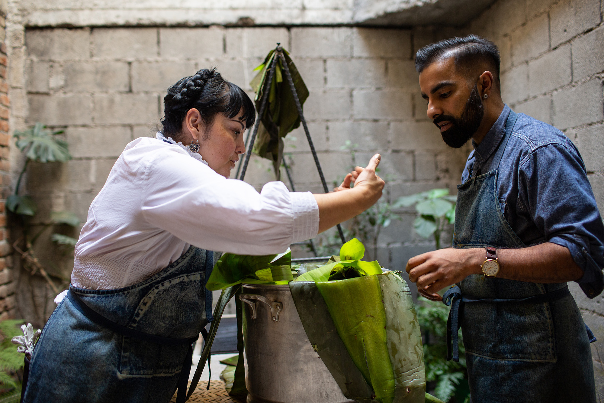 In an outdoor courtyard, a man and woman in blue aprons prepare banana-leaf tamales in a large pot.