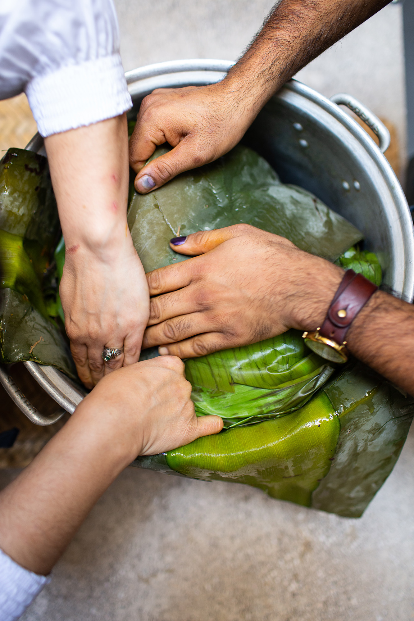 Two pairs of hands pressing down on banana leaves in a large pot.