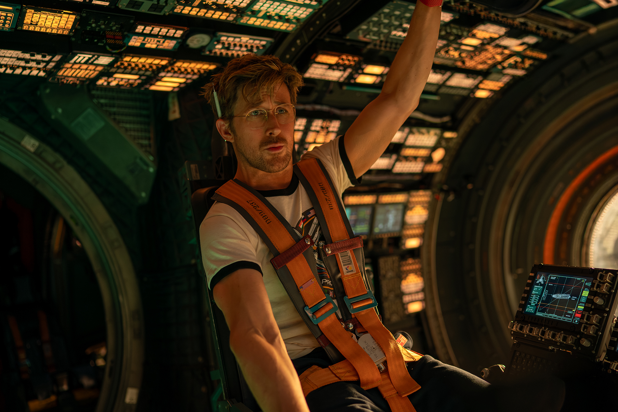 A man in an orange vest and glasses sits inside a spaceship cockpit, surrounded by lights and controls