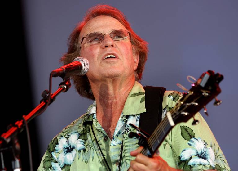 A man in his 70s wears a Hawaiian shirt and glasses while playing an acoustic guitar, singing, and looking upward