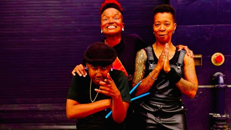 Three Black women in short hair and street clothes look into the camera against a purple wall.