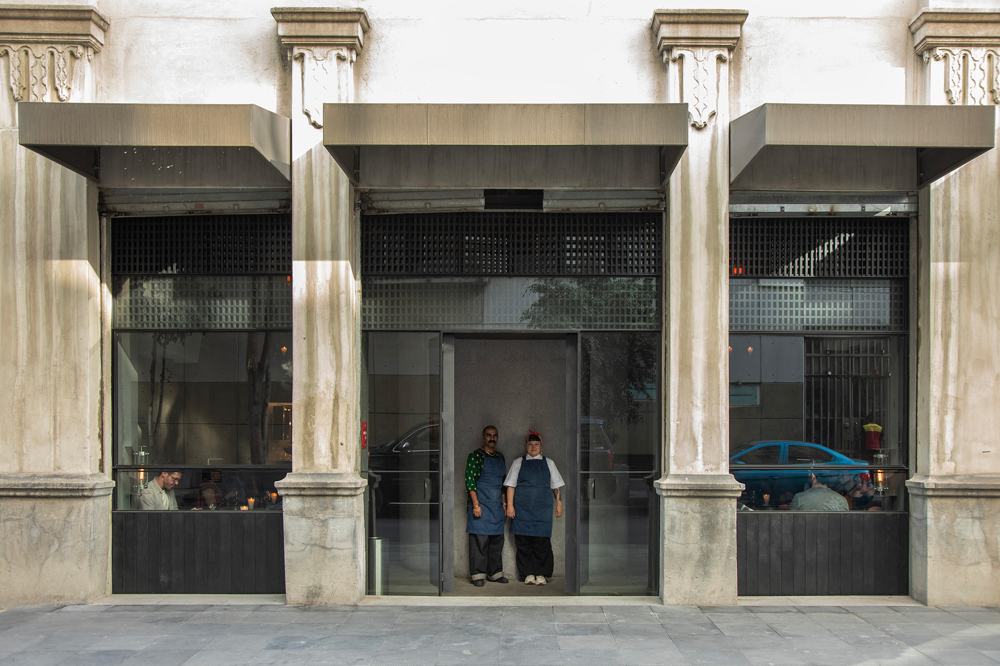 Two chefs in blue aprons pose in front a restaurant, underneath massive cement columns.