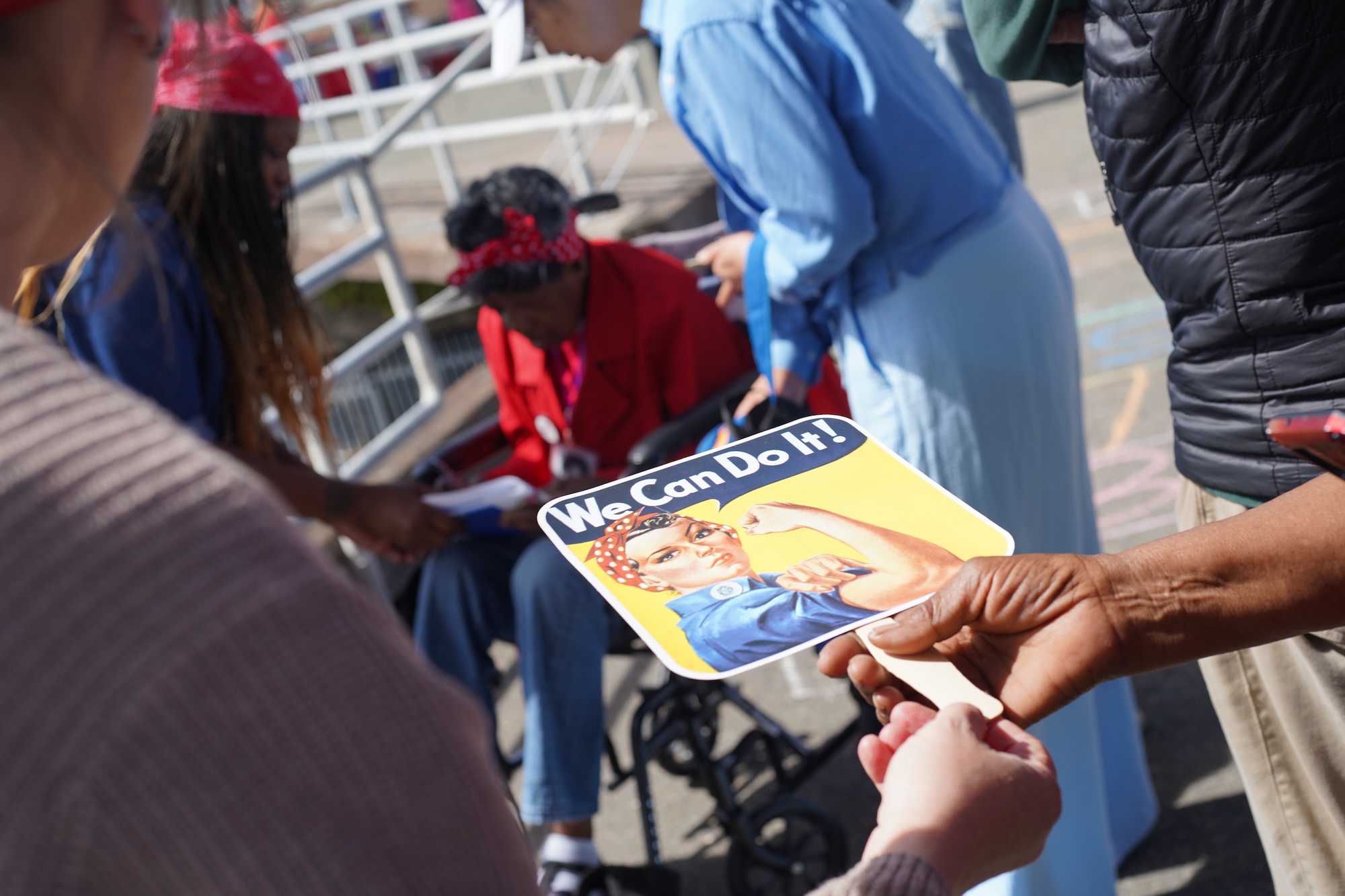 A handheld fan that reads "We Can Do It" is passed in the foreground, as Marcella Hubbard signs an autograph in the background.