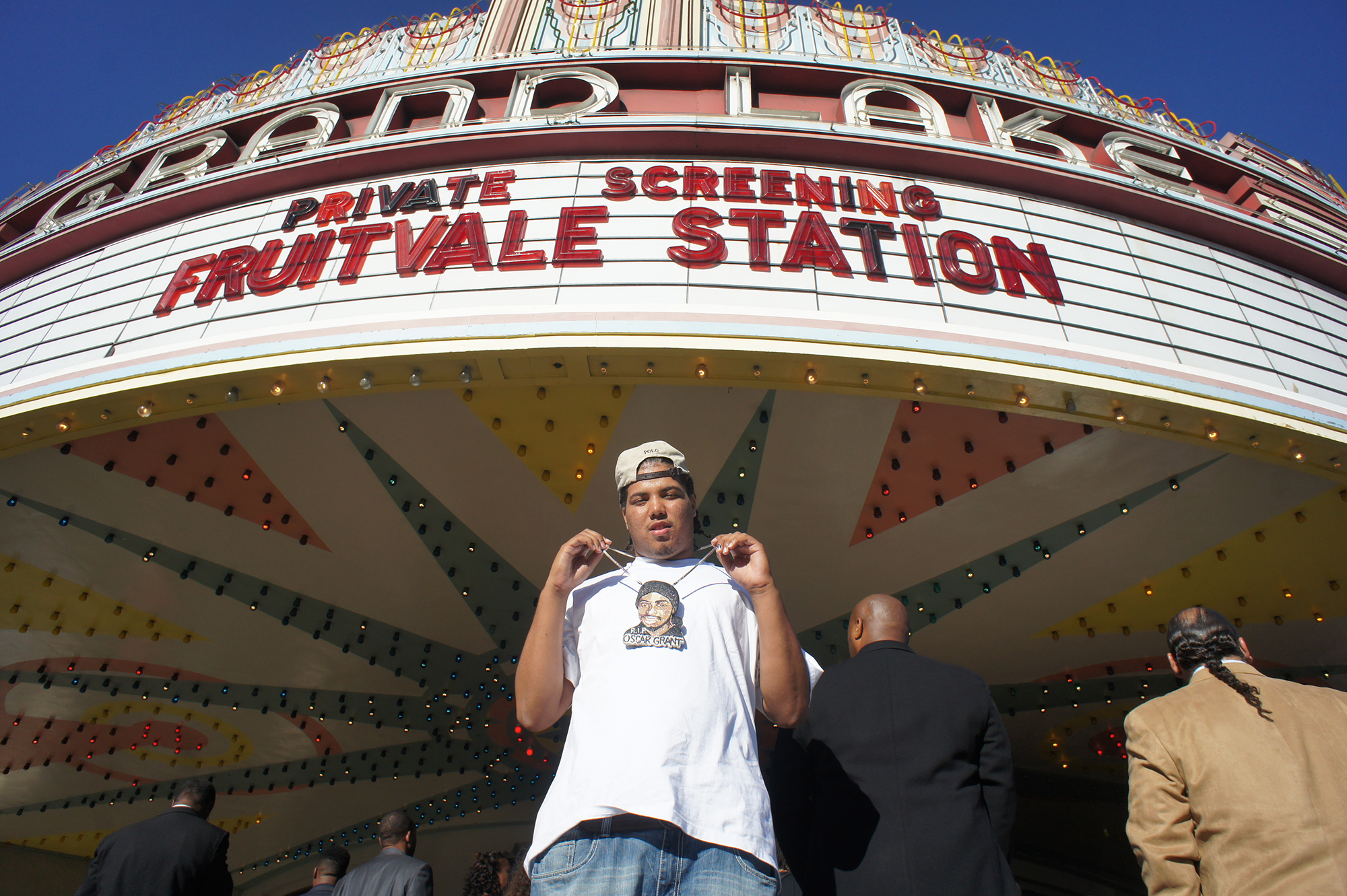 A man in a white cap and T-shirt holds up a medallion in the image of Oscar Grant, beneath a theater marquee reading "Private Screening - Fruitvale Station"