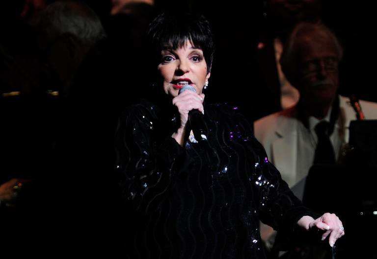 A woman with a black bob haircut and dressed in black sings into a microphone, looking slightly upward