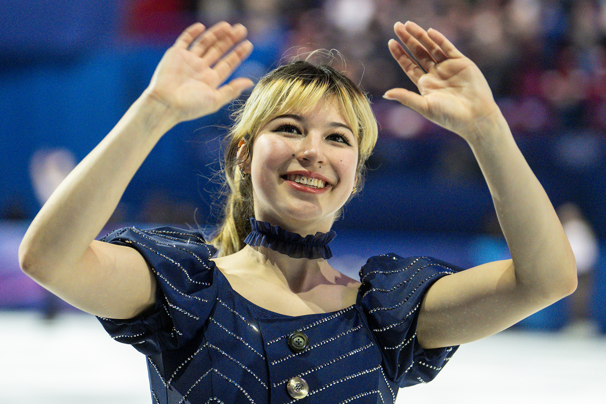 A young woman in a blue outfit and choker necklace waves to a crowd.
