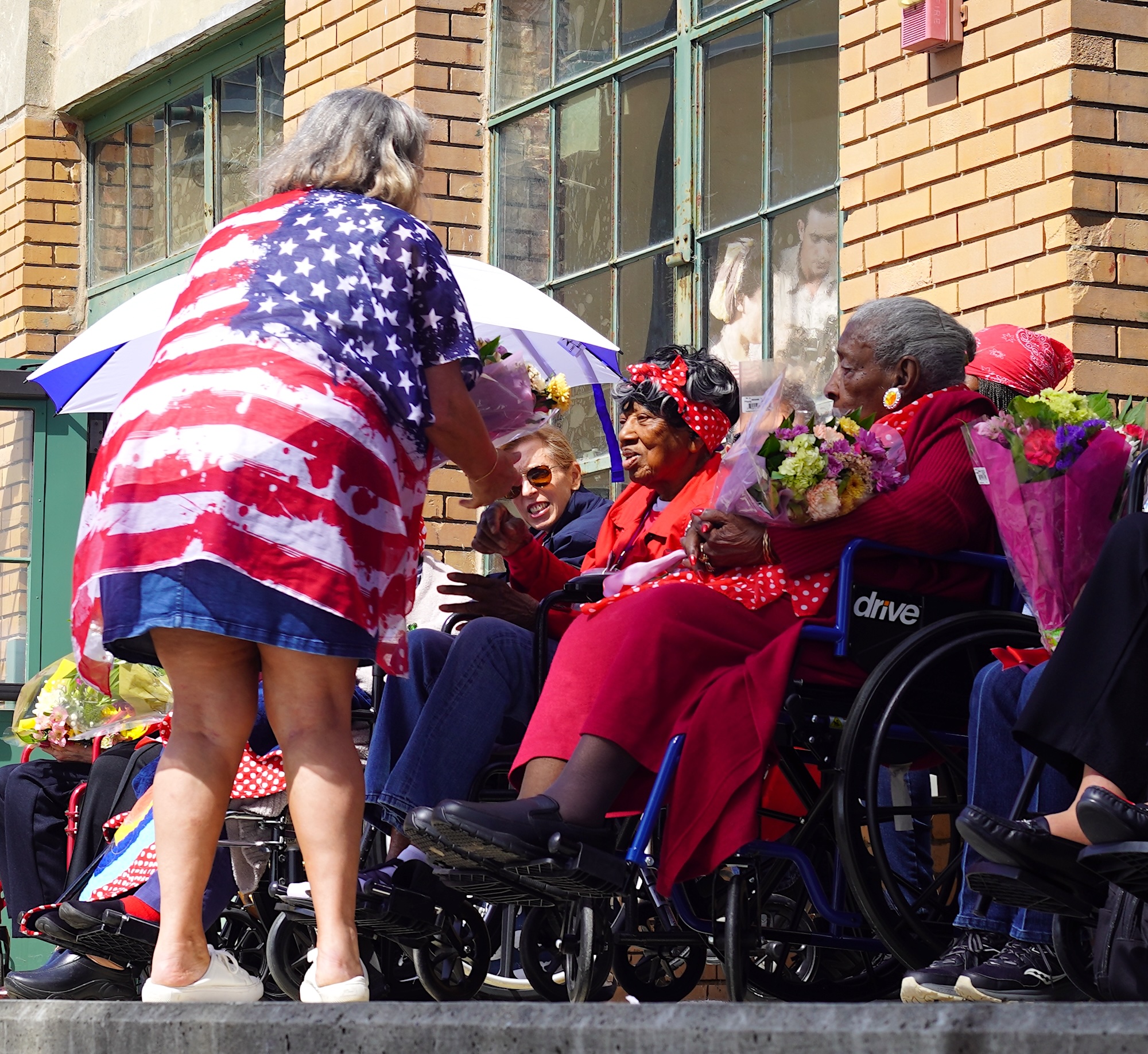 A person in a big USA flag styled shirt passes out flowers to women sitting in wheelchairs on stage. 