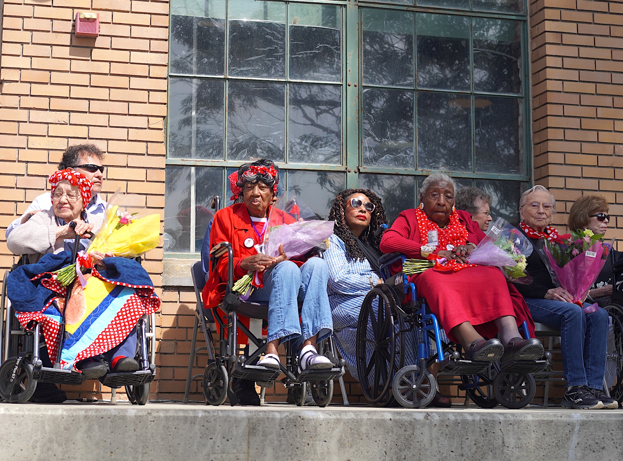 A group of elder women on stage in wheelchairs, holding flowers as they're honored for their service.