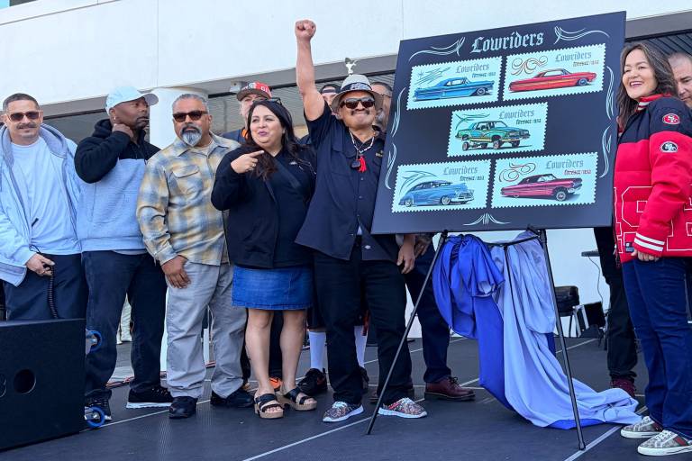 A group of Latino celebrants gather around an easel showing five stamp designs of lowrider-style cars