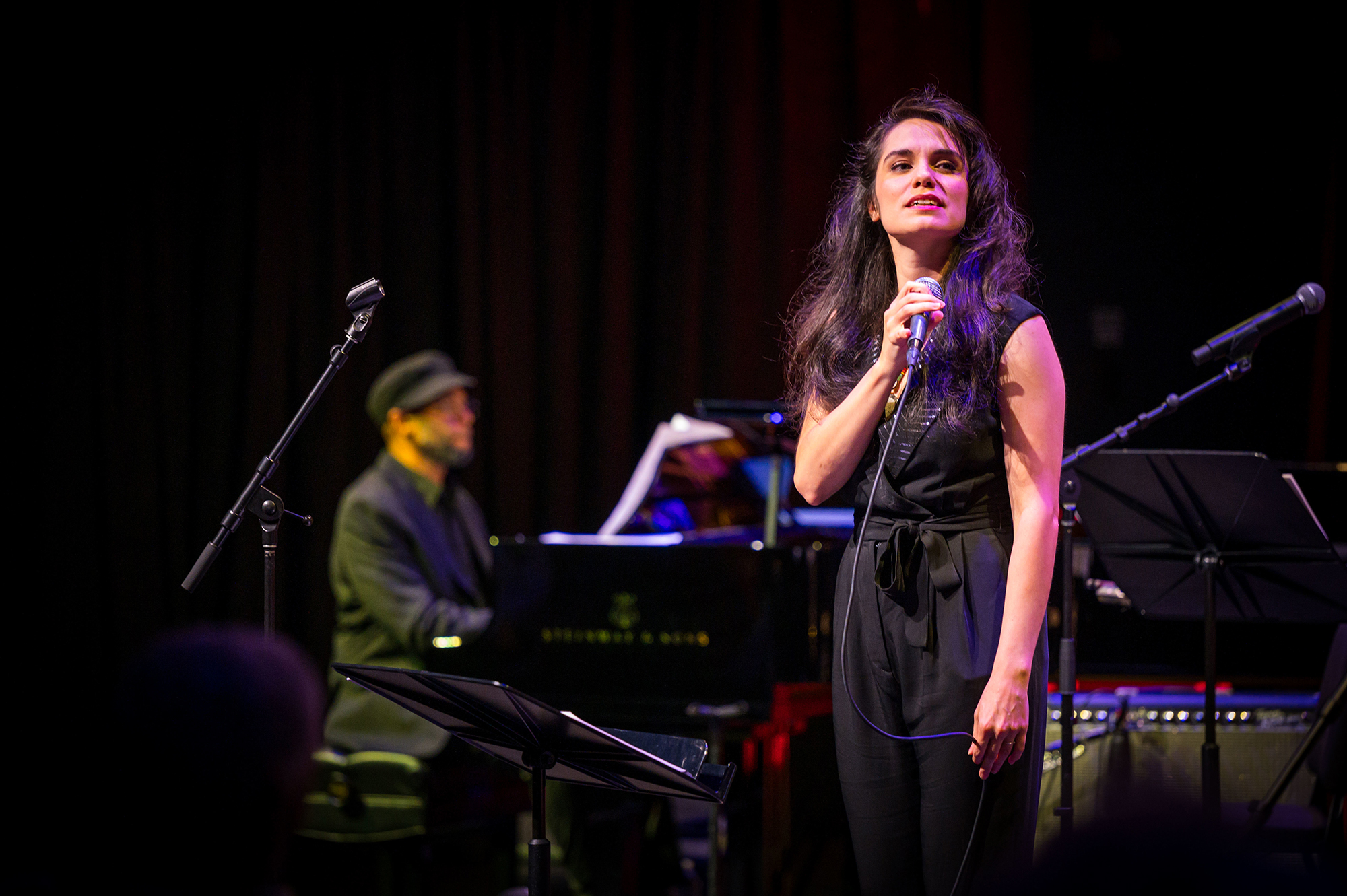 A woman in a black wardrobe holds a microphone and looks slightly upward and to the right, while a pianist plays in the background.