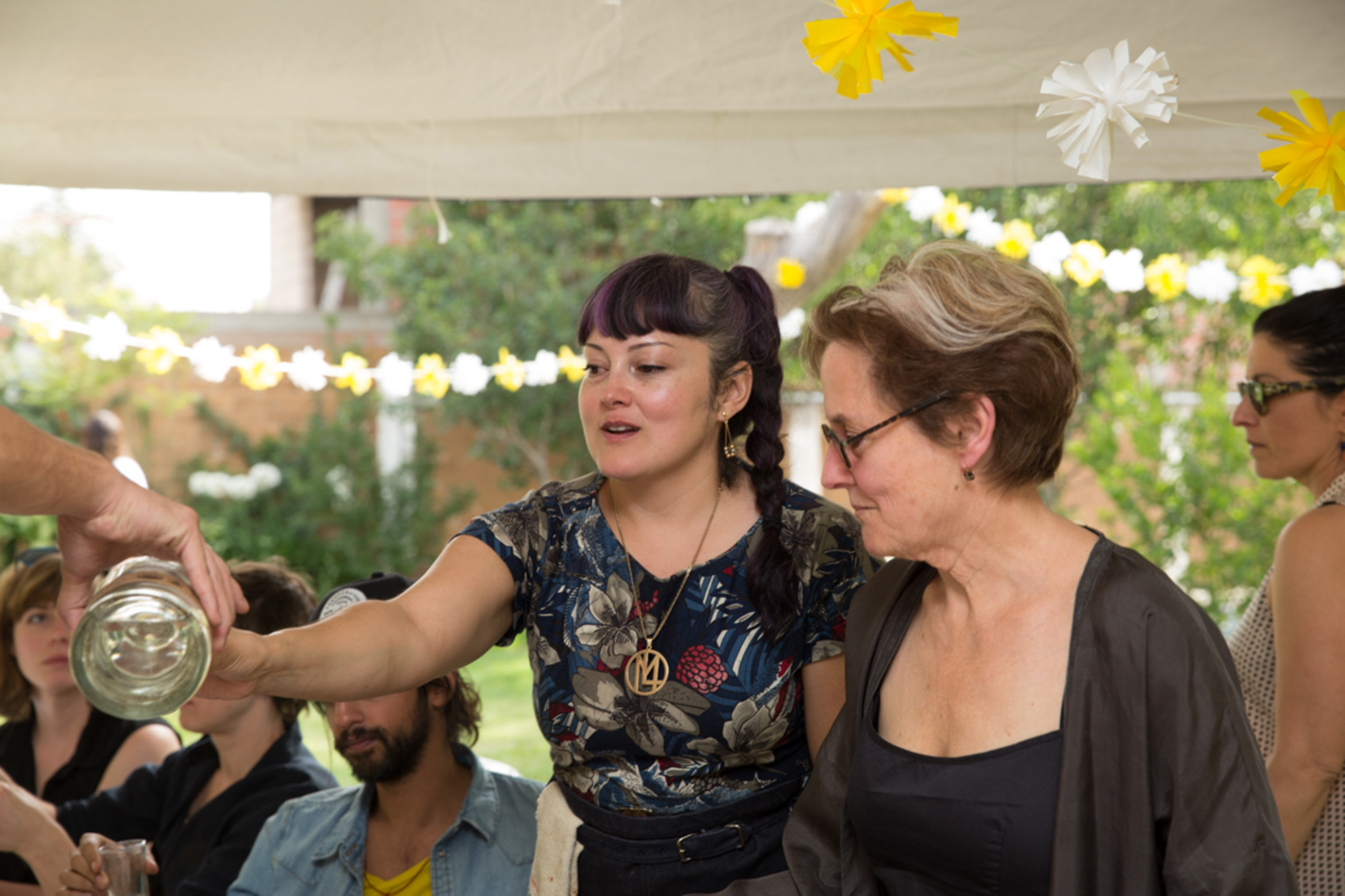 In a backyard garden setting, a woman holds out a glass while a bottle wine is poured.