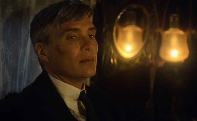 A white middle aged man stands in a dimly lit room, wearing a serious expression.