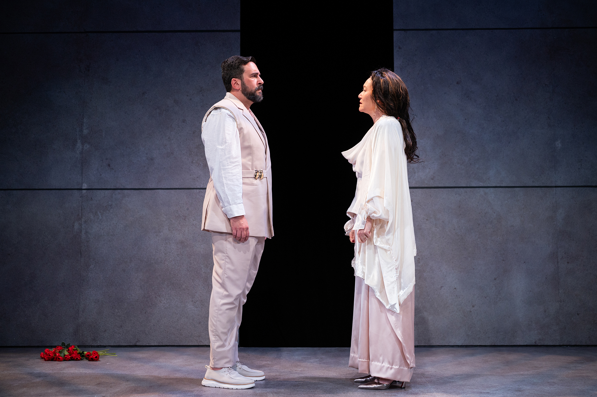 A man ad a woman both dressed in white and cream colors face each other in profile, dramatically, against a black and gray background