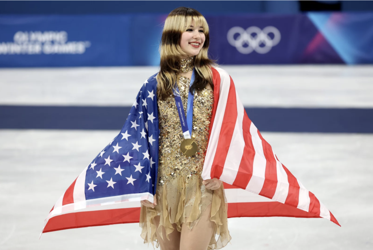 A female figure skater wearing a gold, beaded outfit stands on the ice wrapped in an American flag and wearing a gold medal. She is smiling broadly. Her hair is dyed to have circular black and white stripes.