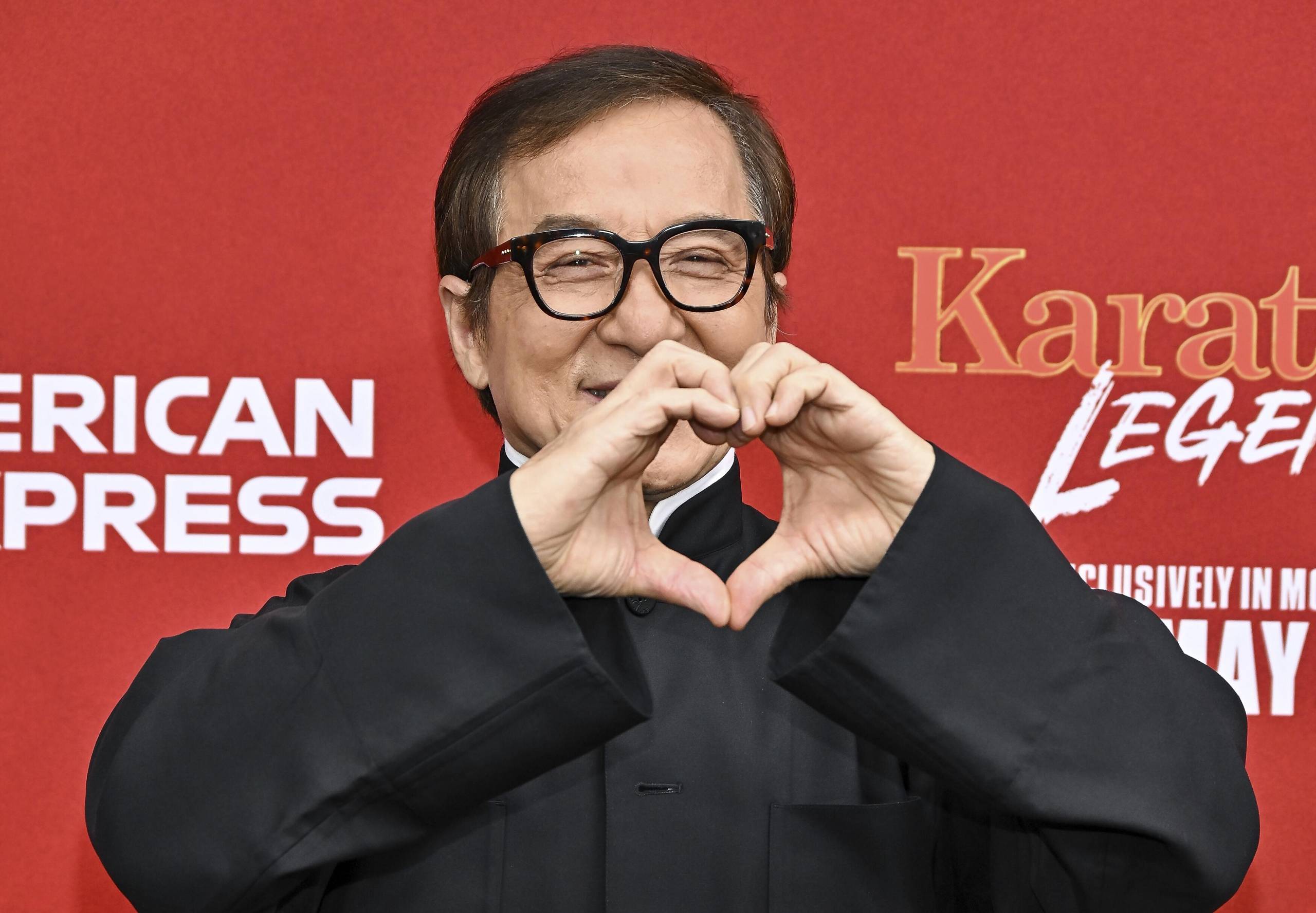 Jackie Chan stands on a red carpet wearing a black suit and spectacles. He is holding up his hands to make a heart shape.