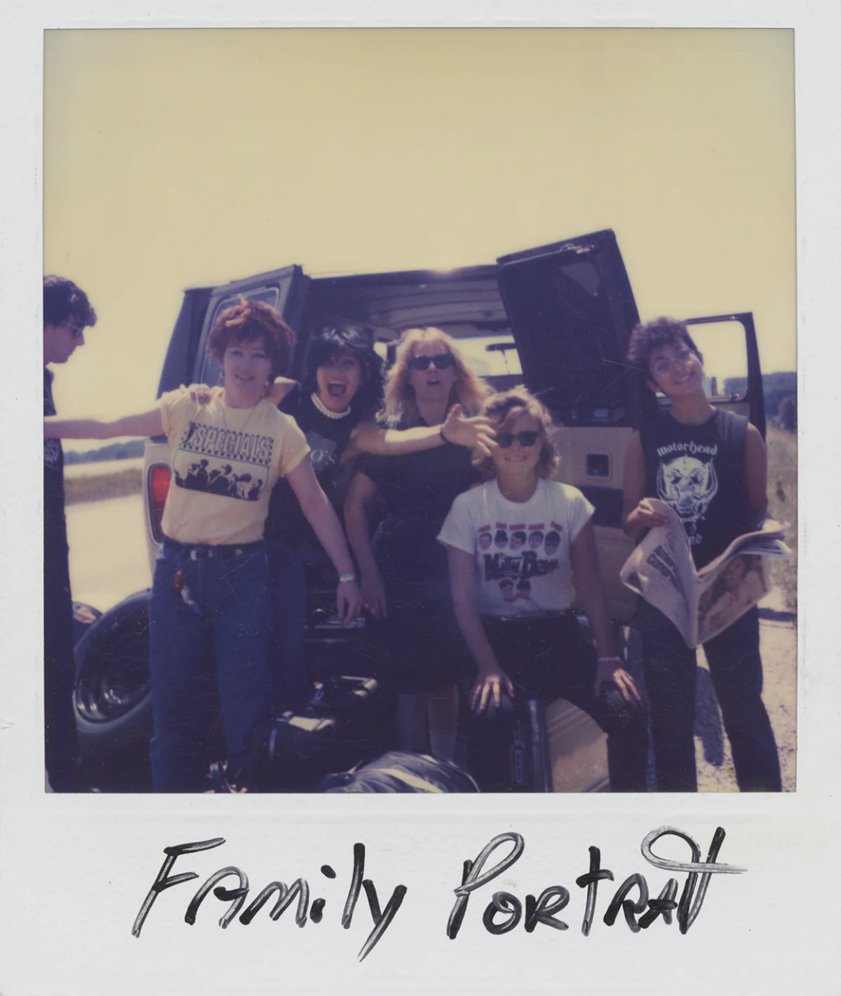 A group of five young women in jeans and band t-shirts, at the back of a black broken down van at the side of the freeway, smile for a Polaroid.