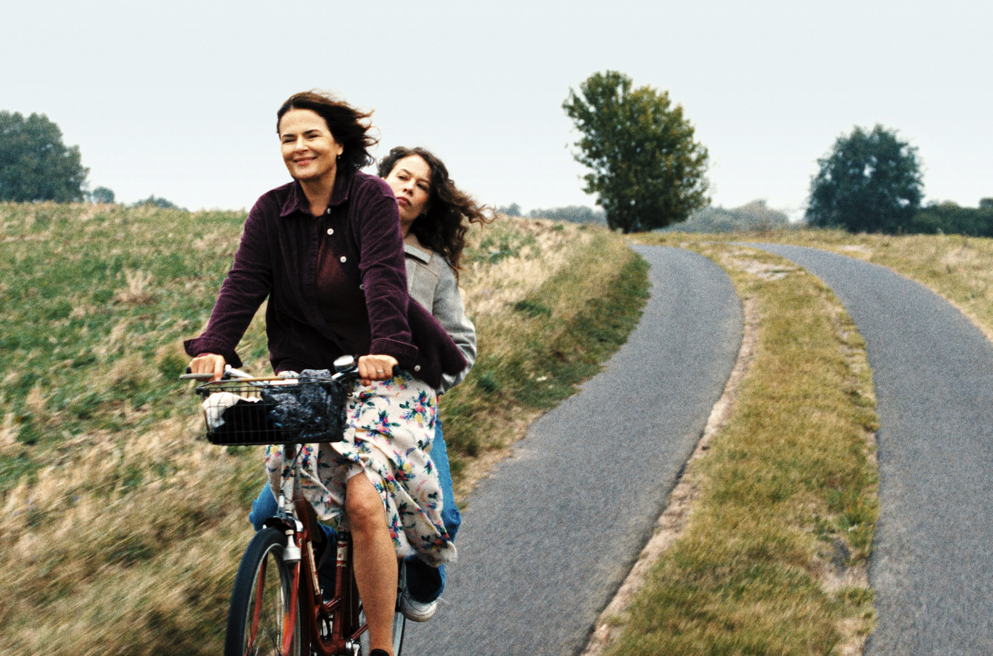 A middle aged white woman happily rides a bicycle through the countryside. A female companion sits behind her, looking stern.