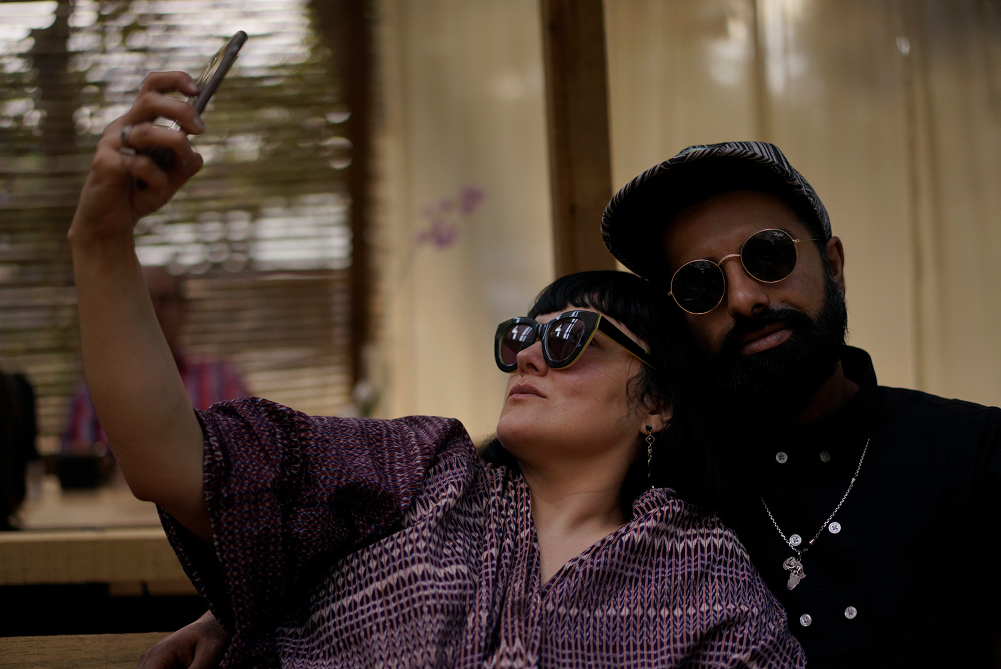A man and a woman, both in sunglasses, sit close together while the woman takes a selfie of the two of them with her phone.
