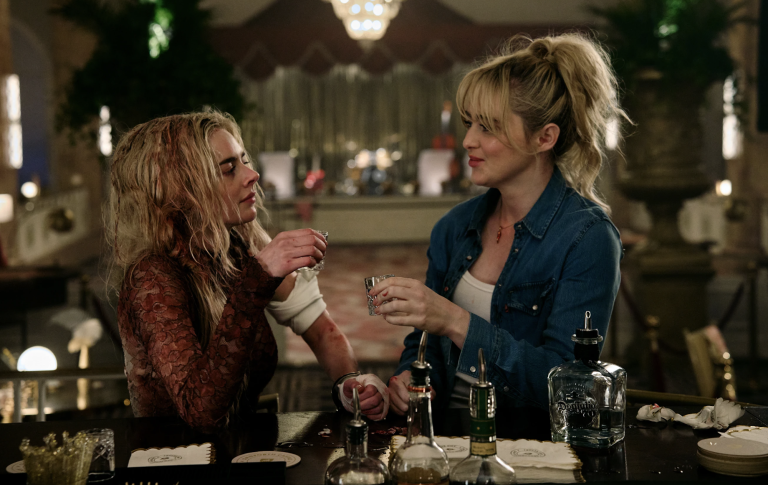 Two 30-something, white blond women sit at a bar in a lavish empty ballroom, casually having a drink. One has matted hair and a blood soaked dress on, the other is dressed casually in a white top and denim shirt.